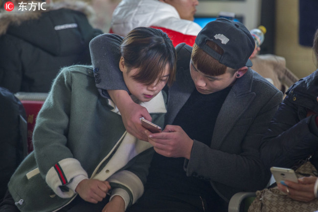 A young man is playing with his smartphone with his girlfriend beside him in a railway station in Hefei. [IC]