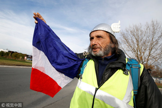 A protesters wearing yellow vest, the symbol of a French drivers' protest against higher diesel fuel prices, holds a French flag as he occupies a roundabout with comrades in Sainte-Eulalie near Bordeaux, France, December 5, 2018. The slogan reads "Macron Get Out". [Photo: VCG/Regis Duvignau]