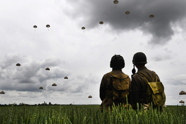 Men dressed as US GIs stand in a field as paratroopers from Britain's 16 Air Assault Brigade and France's 11th Parachute Brigade perform a jump over Sannerville, north-western France, on June 5, 2019, prior to D-Day commemorations marking the 75th anniversary of the World War II Allied landings in Normandy. [Photo: AFP/Fred Tanneau]