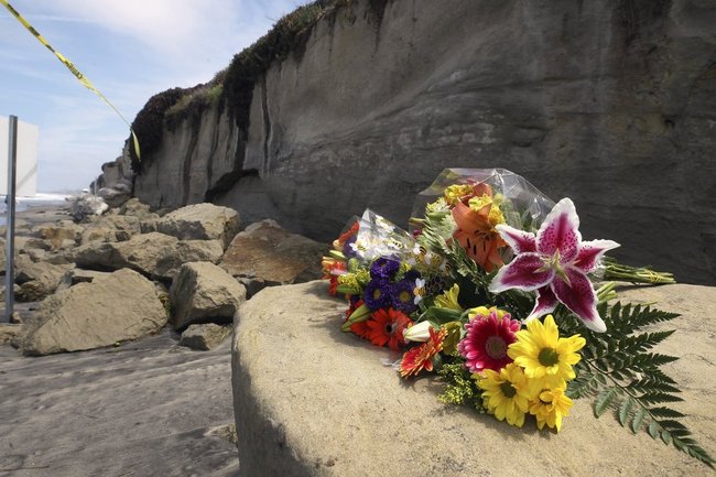 A bouquet of flowers lies on some of the sand rock debris from Friday's bluff collapse, which killed three people, near the Grandview Beach access stairway in the beach community of Leucadia, Saturday, Aug. 3, 2019, in Encinitas, Calif. Officials have reopened much of the Southern California beach where a sea cliff collapsed Friday. Encinitas Lifeguard Capt. Larry Giles said Saturday that a lifeguard will be posted near the collapse zone on Grandview Beach, which is still marked by yellow caution tape. [Photo: AP/Hayne Palmour IV/The San Diego Union-Tribune]