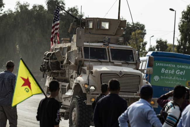 Syrian Kurds, one of them carrying a Kurdish YPG (People's Protection Units) flag, watch as a US military vehicle drives on a road after US forces pulled out of their base in the Northern Syriain town of Tal Tamr, on October 20, 2019. [Photo: AFP/Delil Souleiman]