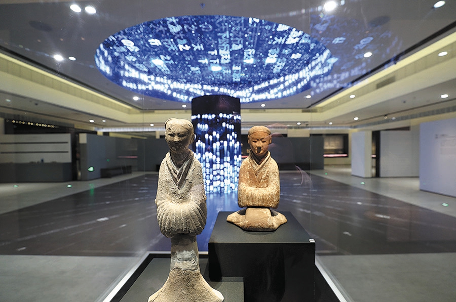 Δύο κεραμικά αγαλματίδια στο φόντο μιας συσκευής πολυμέσων, η οποία απεικονίζει τη μεταμόρφωση των κινεζικών χαρακτήρων. [Φωτογραφία ZOU HONG/CHINA DAILY]